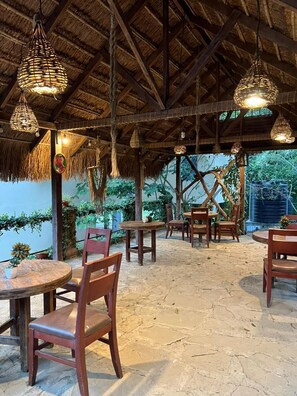Un área de comedor al aire libre con mesas y sillas de madera, techo de paja, lámparas colgantes y follaje en el fondo.