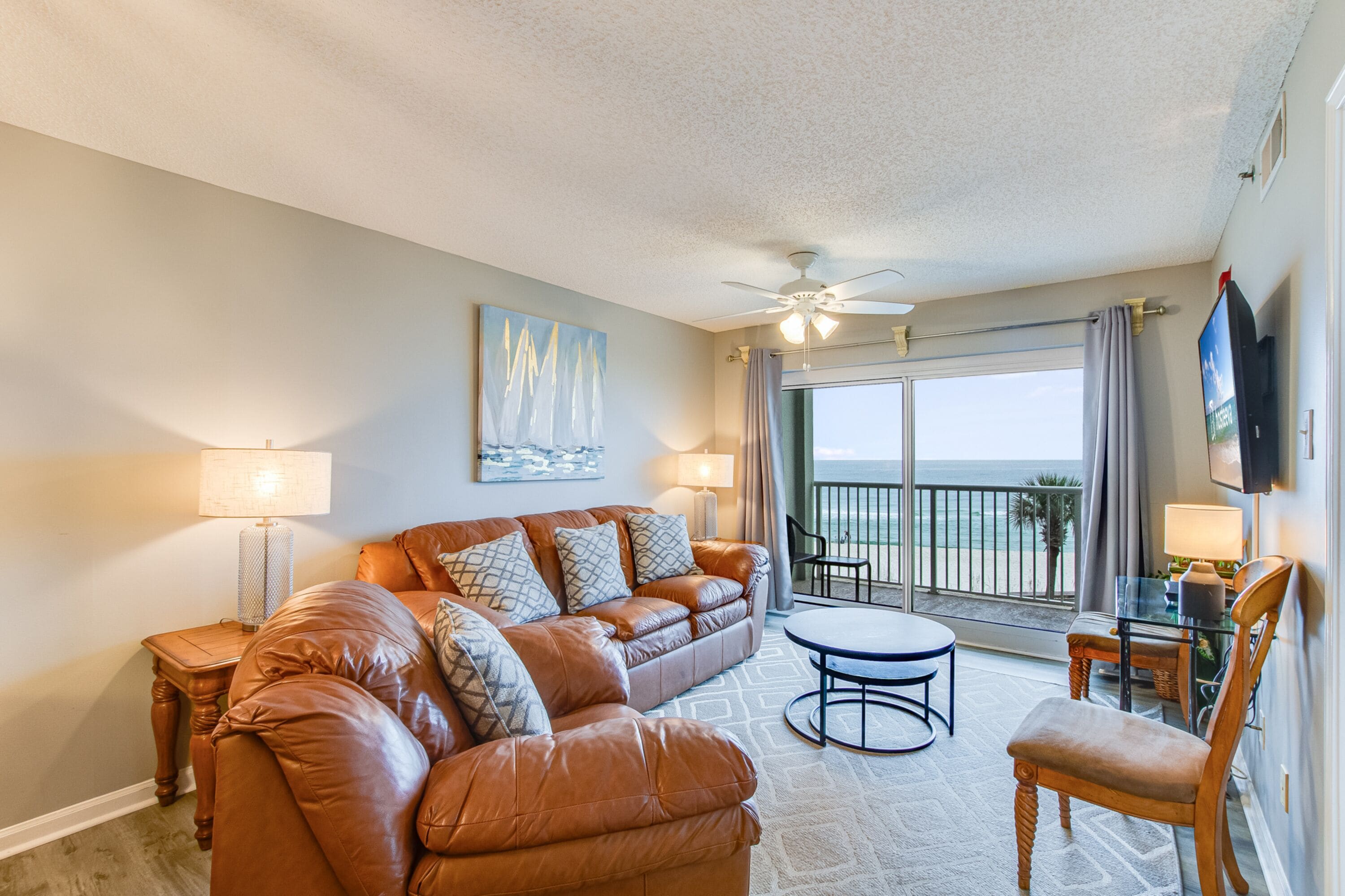 A living room with a brown leather sofa, a glass coffee table, and a TV on the wall. There is a balcony with a view of the ocean.