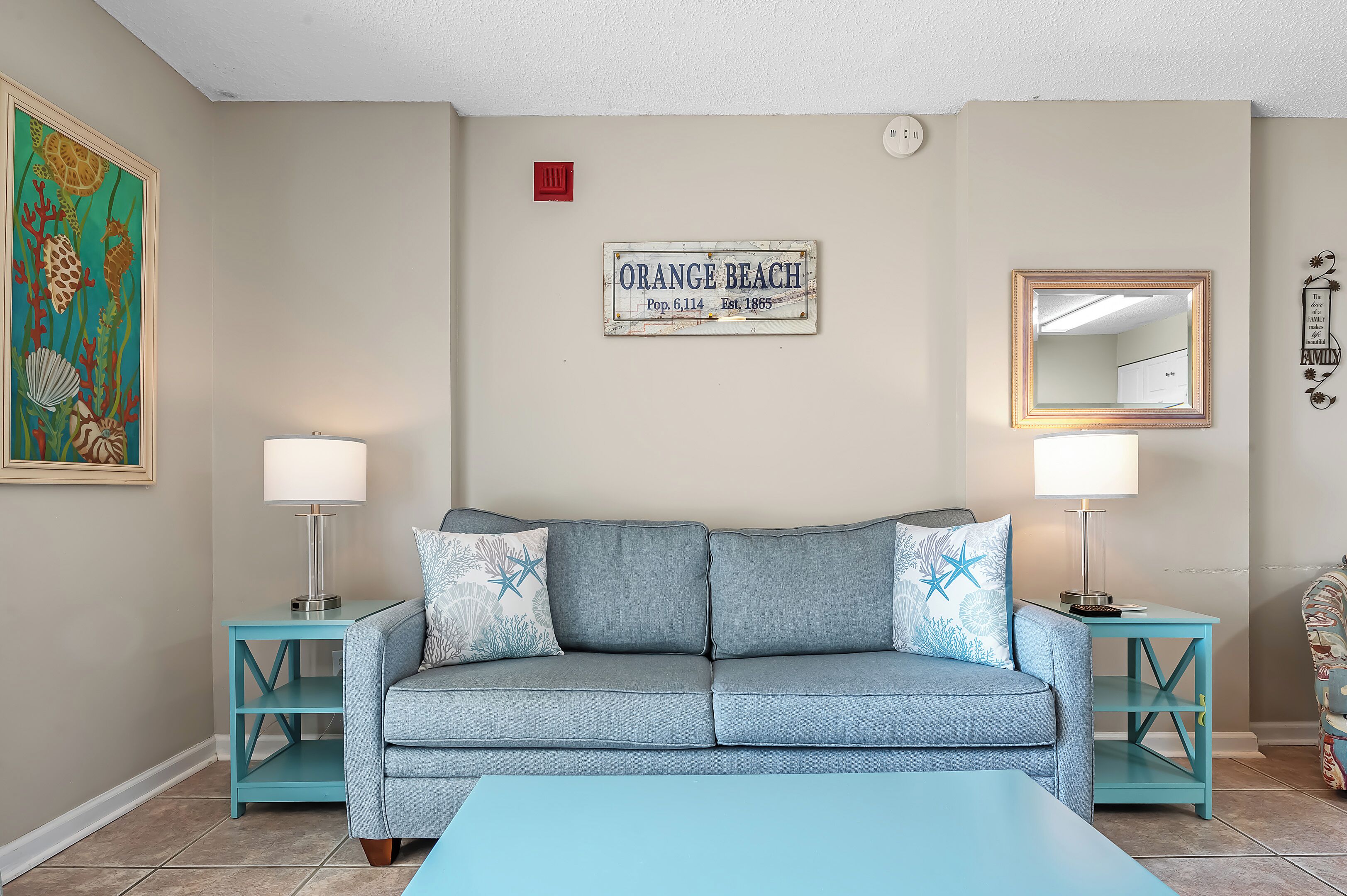 A living room with a light blue sofa, two matching side tables, a coffee table, a framed sign on the wall, a painting, and a mirror.