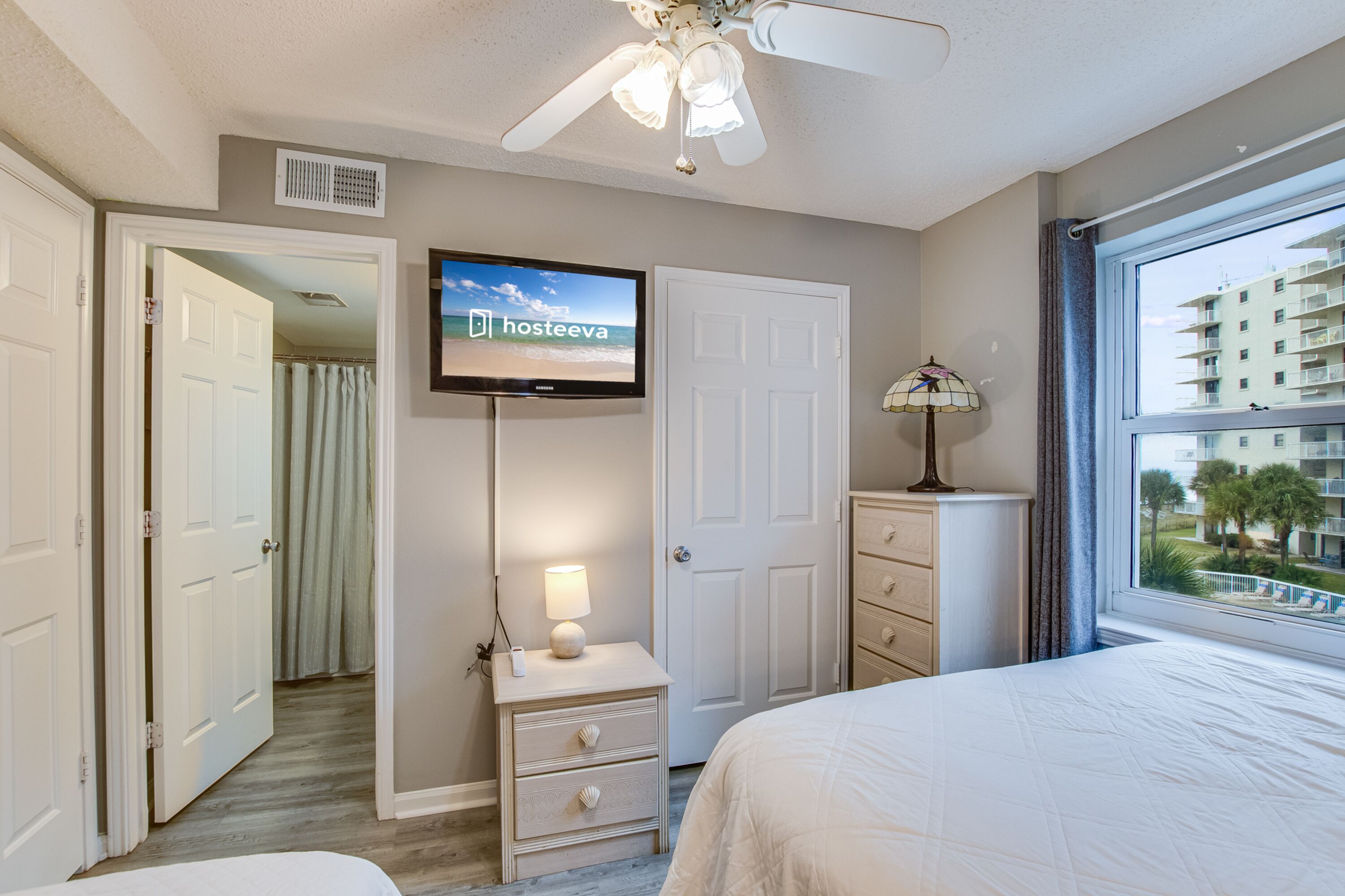 A hotel room with a bed, a TV mounted on the wall, a ceiling fan, a nightstand with a lamp, and a view of the outside through a window.