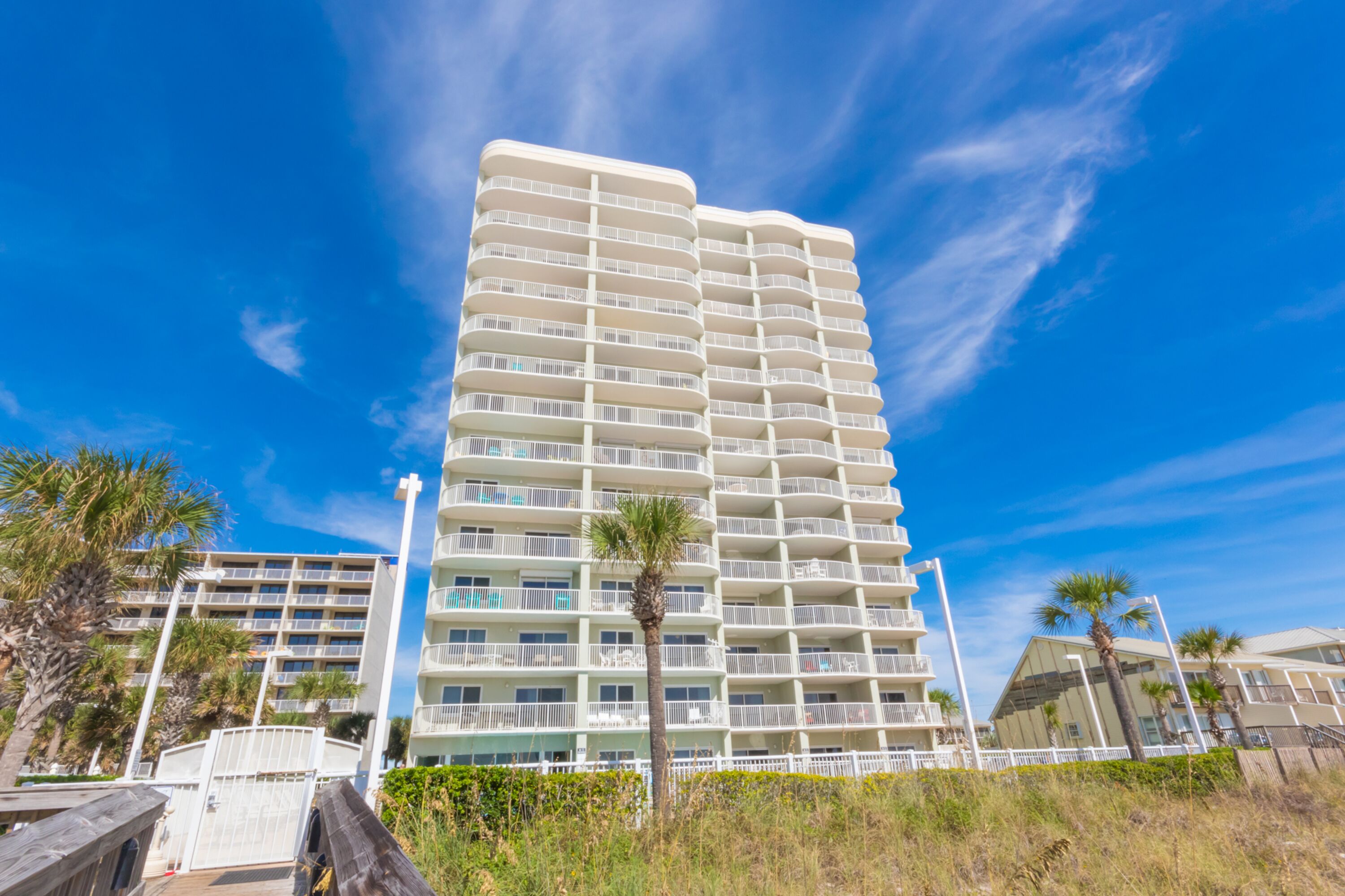 Un grand hôtel à plusieurs étages, doté de balcons, situé près d’une plage bordée de palmiers, sous un ciel bleu azur.