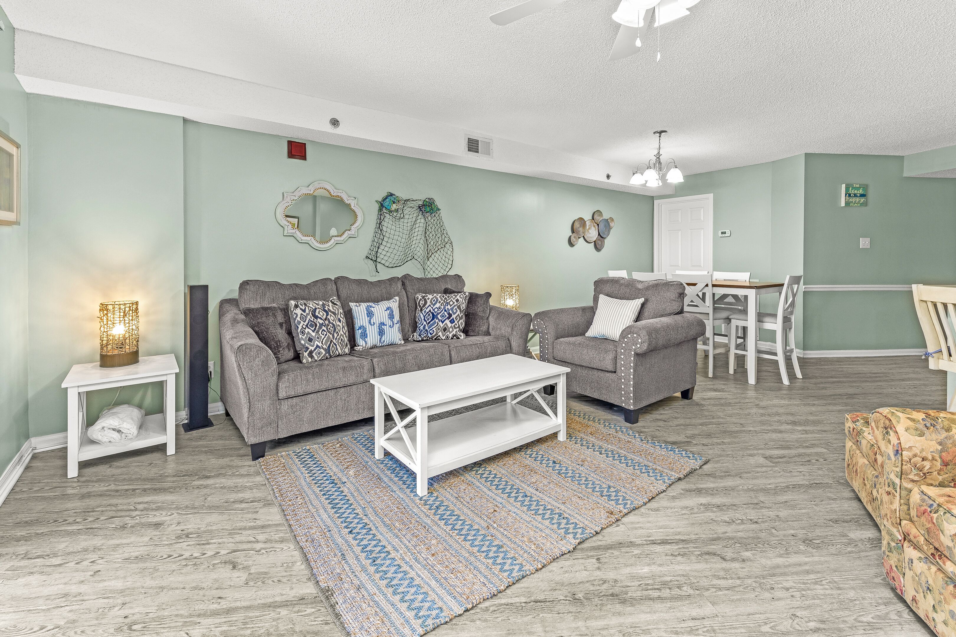 A living room with two grey sofas, a white coffee table, a patterned rug, and a dining area with a white table and chairs.