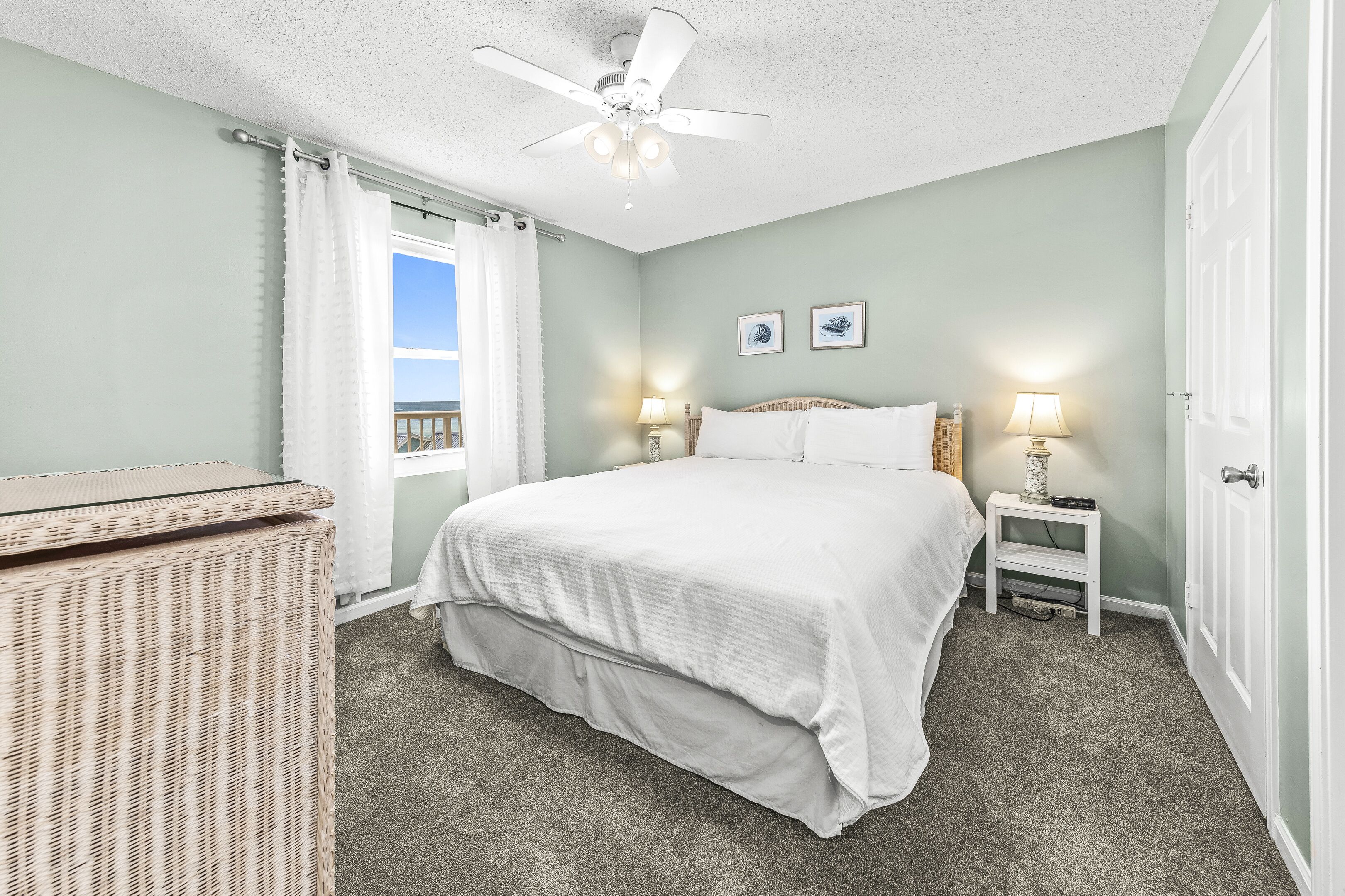 A bedroom with a bed, two bedside lamps, a ceiling fan, a wicker side table, and a window with curtains.