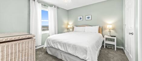 A bedroom with a bed, two bedside lamps, a ceiling fan, a wicker side table, and a window with curtains.