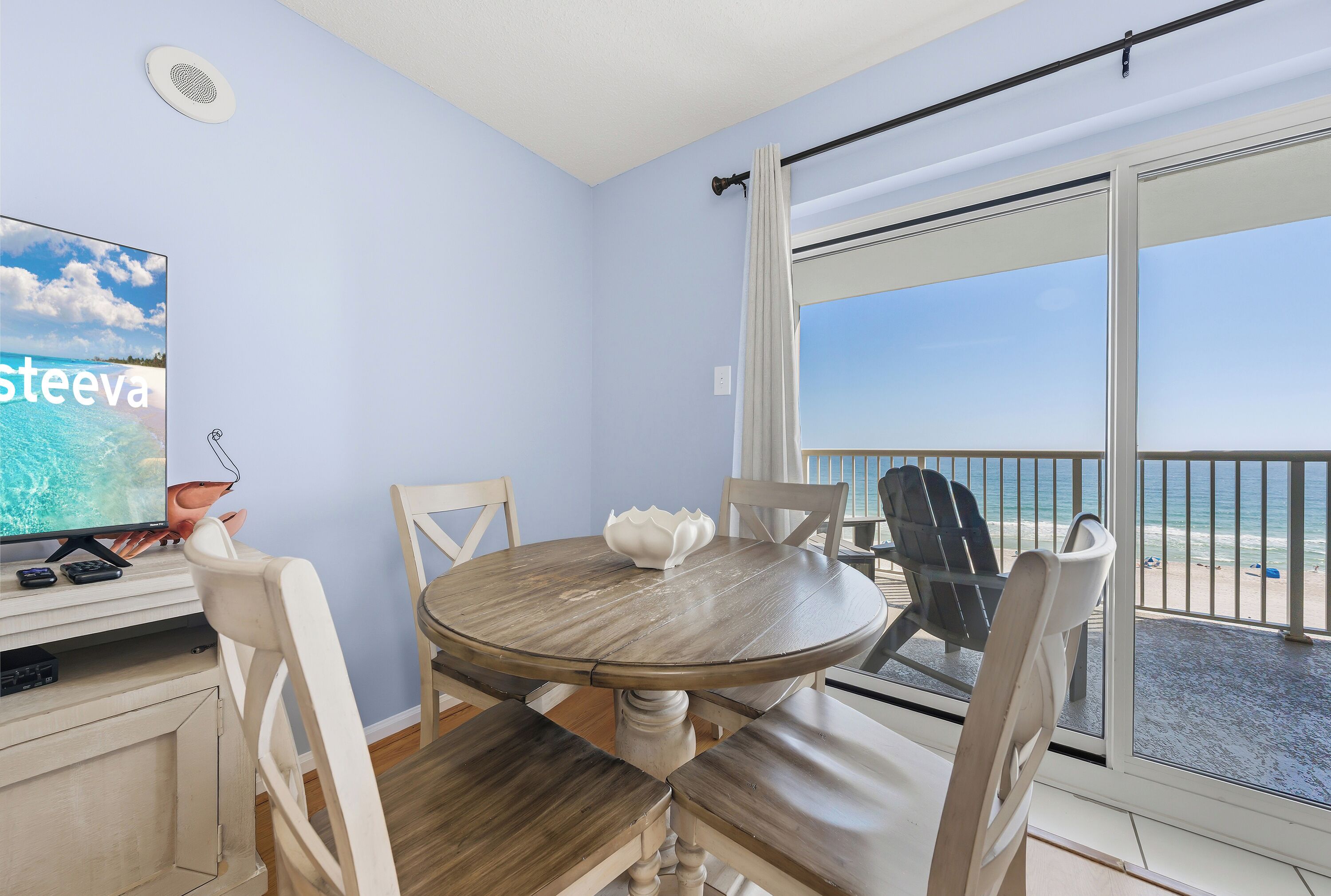 A room with a round wooden table, four chairs, a television, and a view of the beach through a sliding glass door.