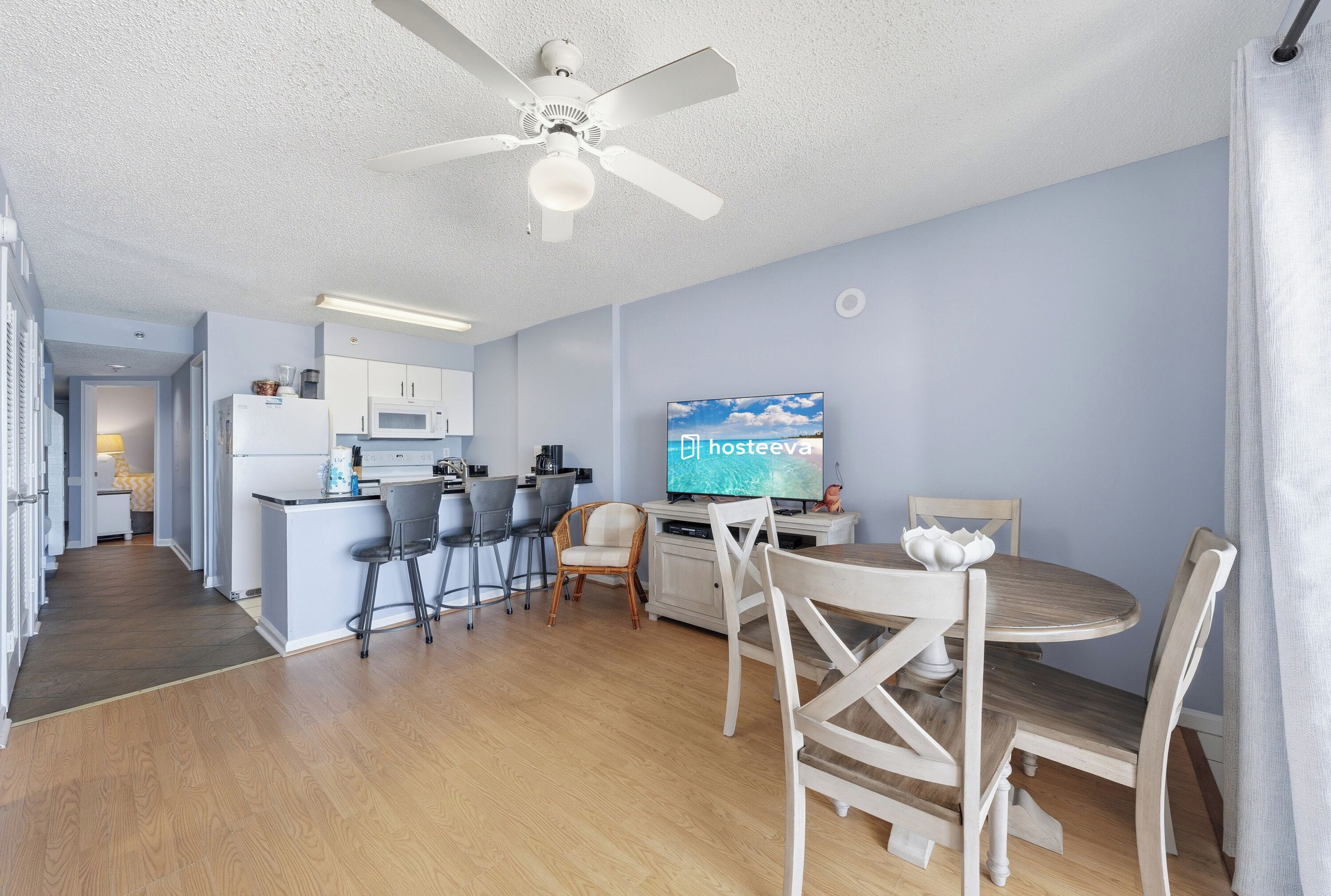 A modern kitchen with a microwave, refrigerator, and bar area. There is a dining table with chairs and a television displaying 'hosteeva' on the screen.