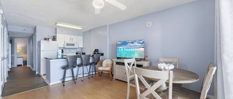 A modern kitchen with a microwave, refrigerator, and bar area. There is a dining table with chairs and a television displaying 'hosteeva' on the screen.