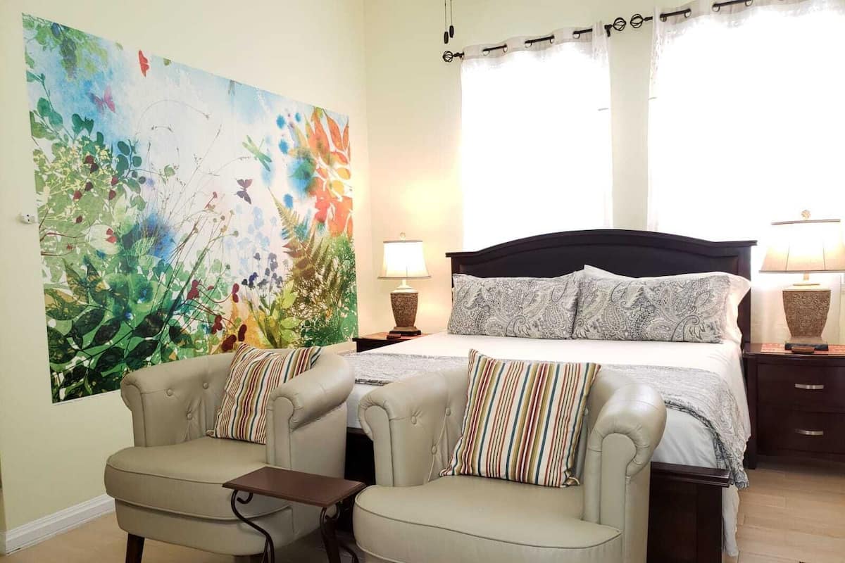 A bedroom with a large floral painting, a bed with patterned pillows, two armchairs, a small table, and two bedside lamps.