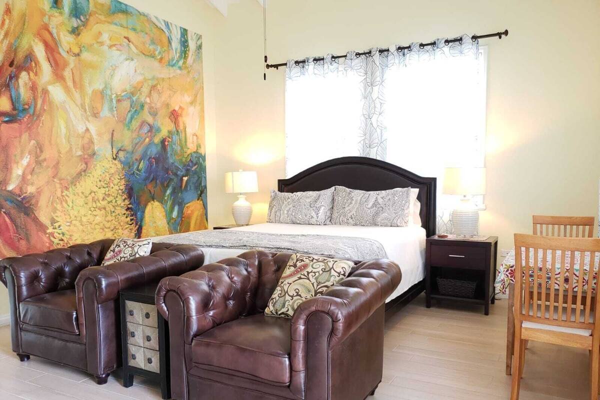 A bedroom with a large bed, two leather sofas, a wooden chair, and a colorful abstract painting on the wall.