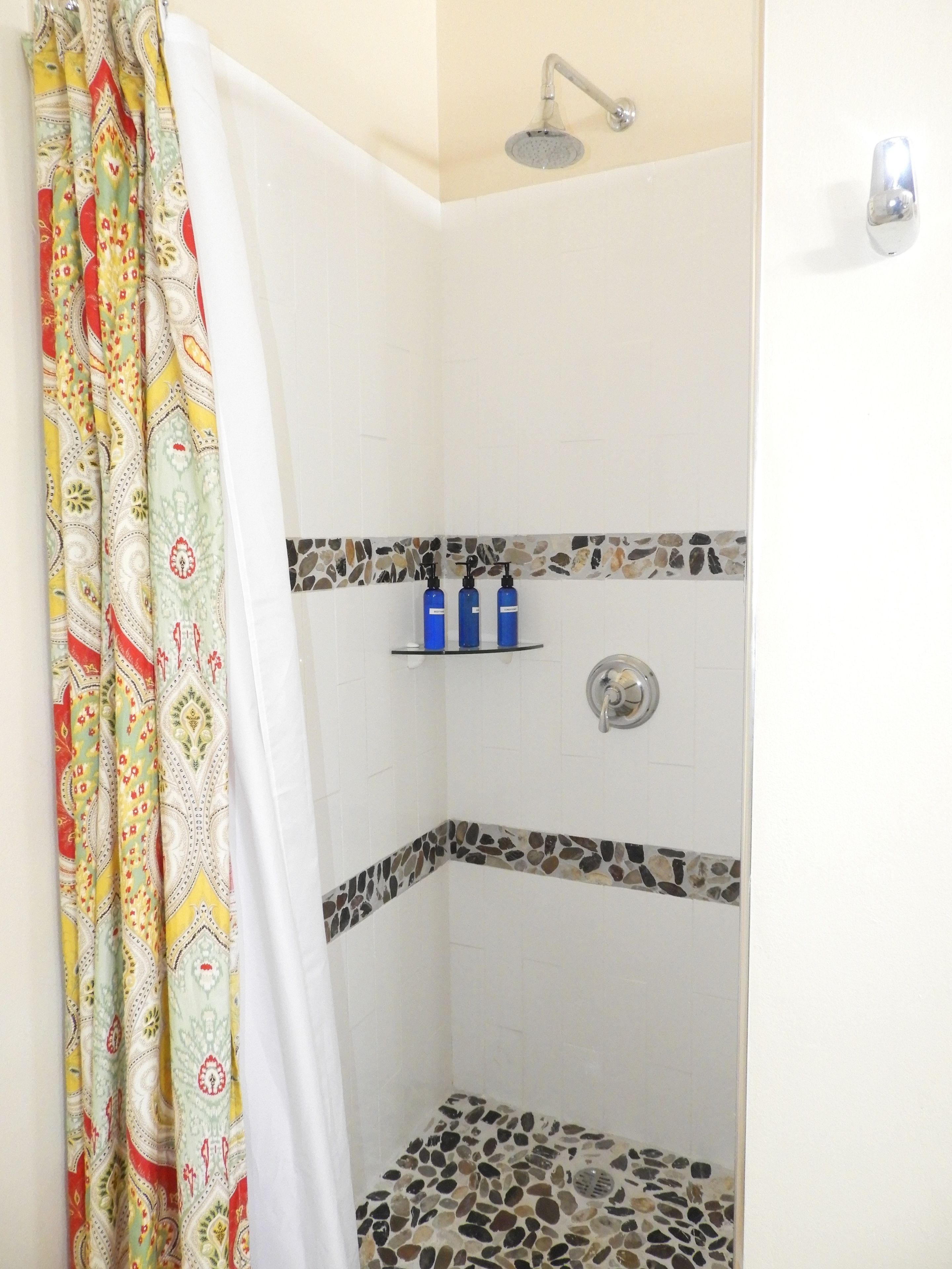 A shower with a rain showerhead, a tiled floor with pebble accents, and a shower curtain with a colorful pattern.