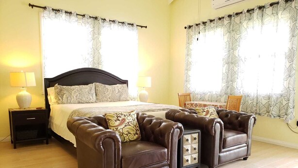 Un dormitorio con una cama, dos sillones de cuero marrón, una mesita de noche, una lámpara y un ventanal con cortinas estampadas.