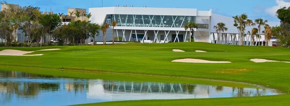 A golf course with a modern building in the background, a pond, and palm trees. - Hotel Turquesa (Playa del Carmen)
