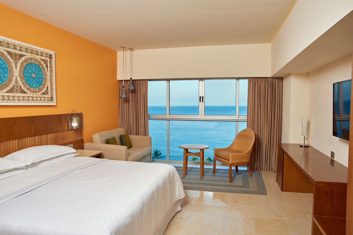Habitación de hotel con cama, sofá, sillón, mesita, televisor y ventana grande con vista al mar.