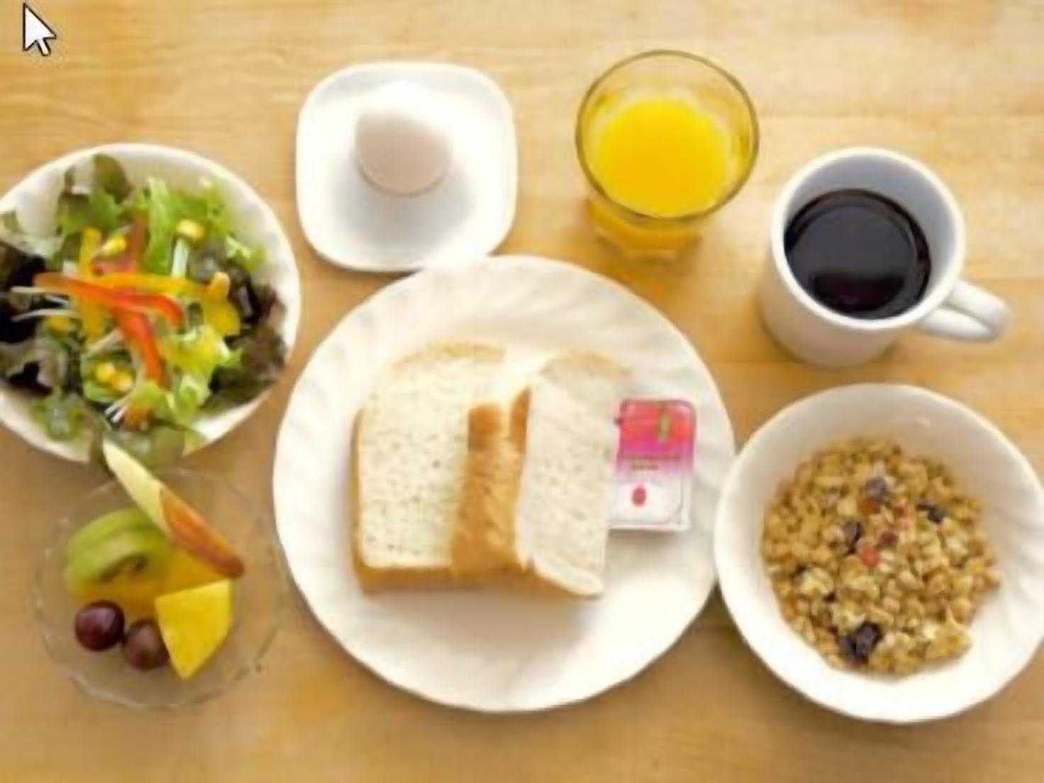 Une table dressée pour le petit-déjeuner avec une assiette de toasts, un bol de céréales, un verre de jus d’orange, une tasse de café, une salade et un panier de fruits.