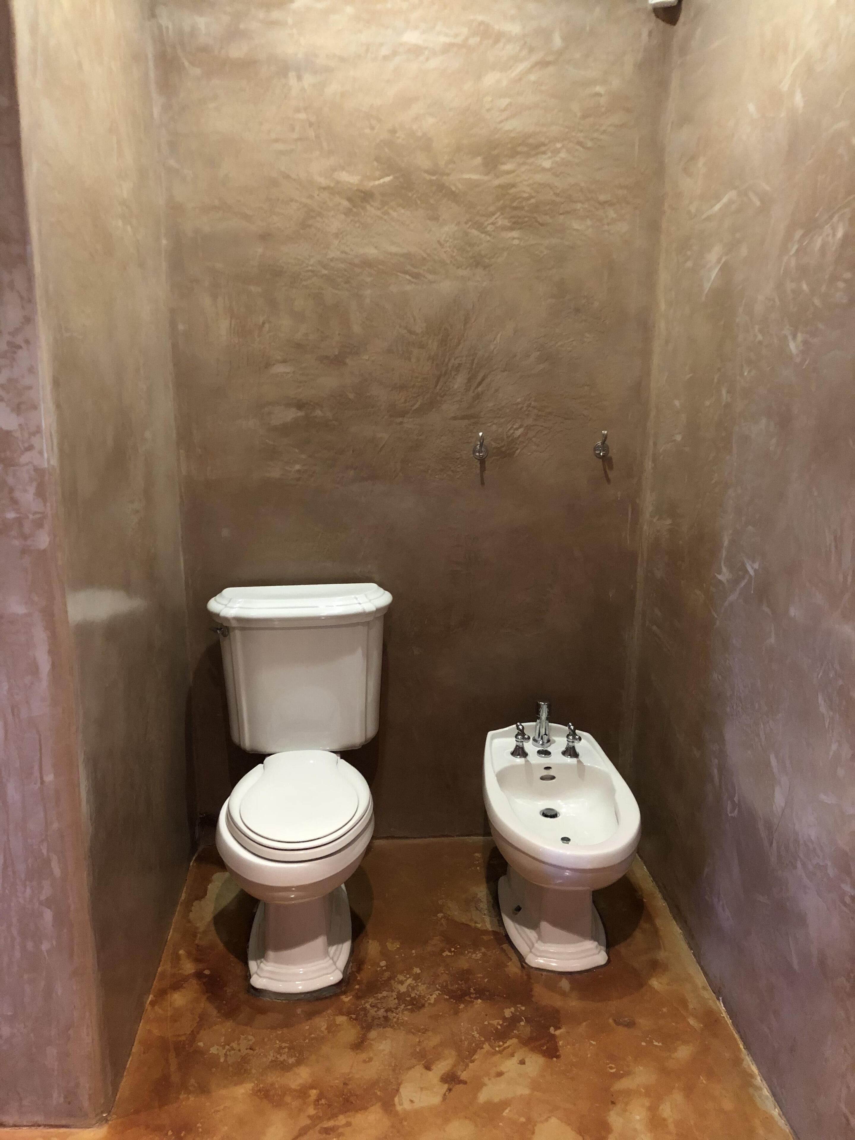 A small bathroom with a toilet and bidet, both in white, against a textured brown wall.