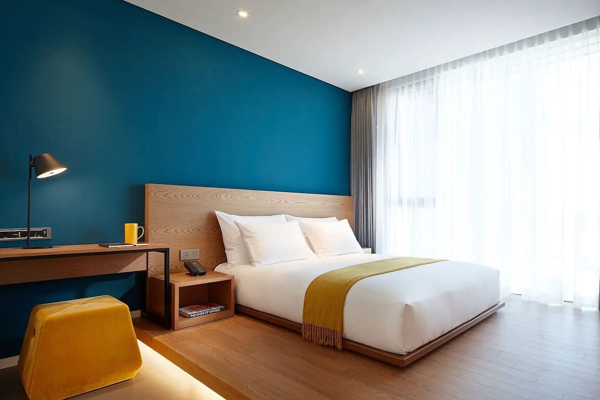 A modern hotel room with a bed, a desk, a chair, a lamp, and a large window with curtains.
