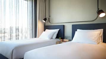 A hotel room with two beds, a nightstand, a lamp, and a view of the city through large windows.