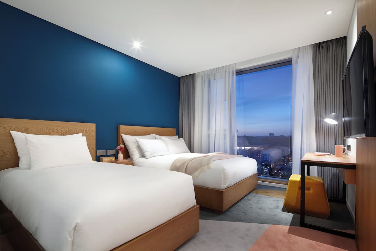 A hotel room with two beds, a desk, a chair, and a large window with a city view.