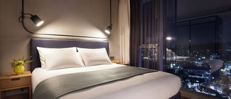 A modern hotel room with a large bed, bedside lamps, a view of the city at night, and a window with curtains.