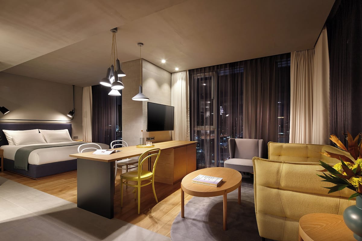 A modern hotel room with a bed, a desk, a chair, a sofa, and a view of the city at night.