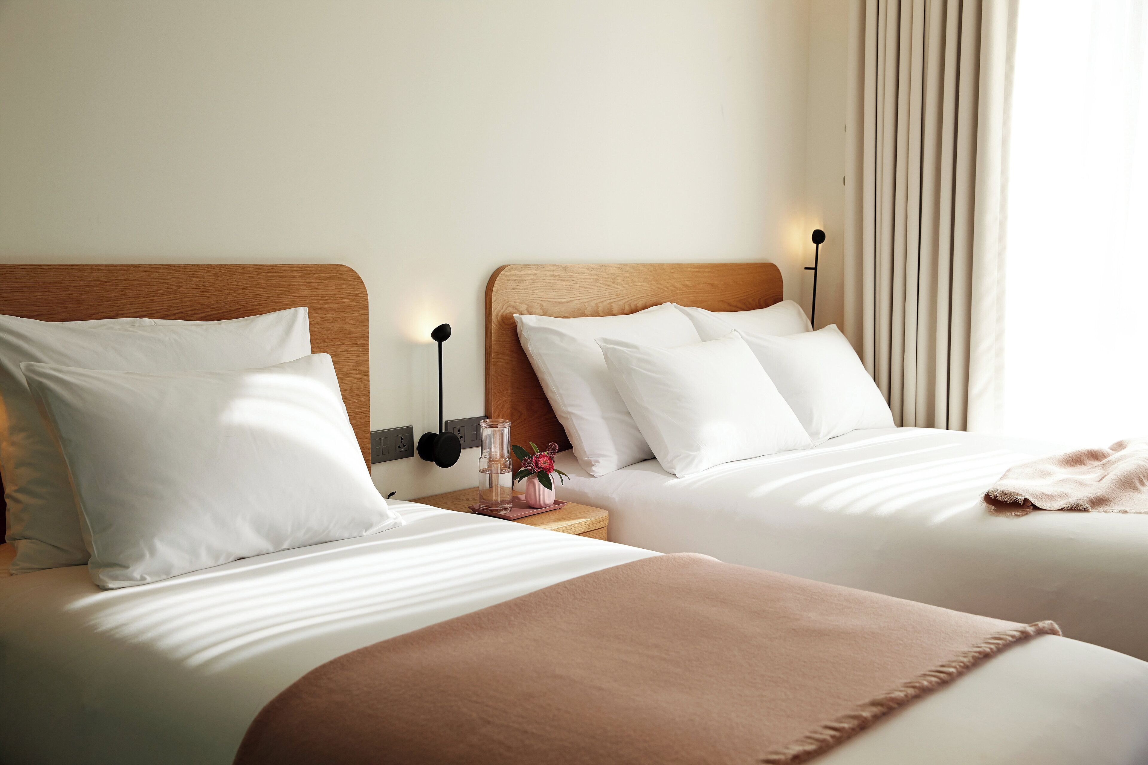 Habitación de hotel con dos camas, cada una con ropa blanca y cabeceros de madera. Hay una mesita de noche con una lámpara y un jarrón con flores.