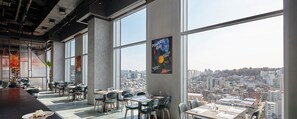 Un espace de restauration moderne doté de grandes fenêtres offrant une vue sur la ville, de tables accompagnées de chaises et d’une affiche accrochée au mur.