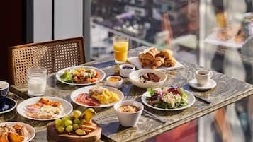 Una mesa de desayuno con diversos platos, como cruasanes, huevos y fruta, en un balcón con vistas a la ciudad.