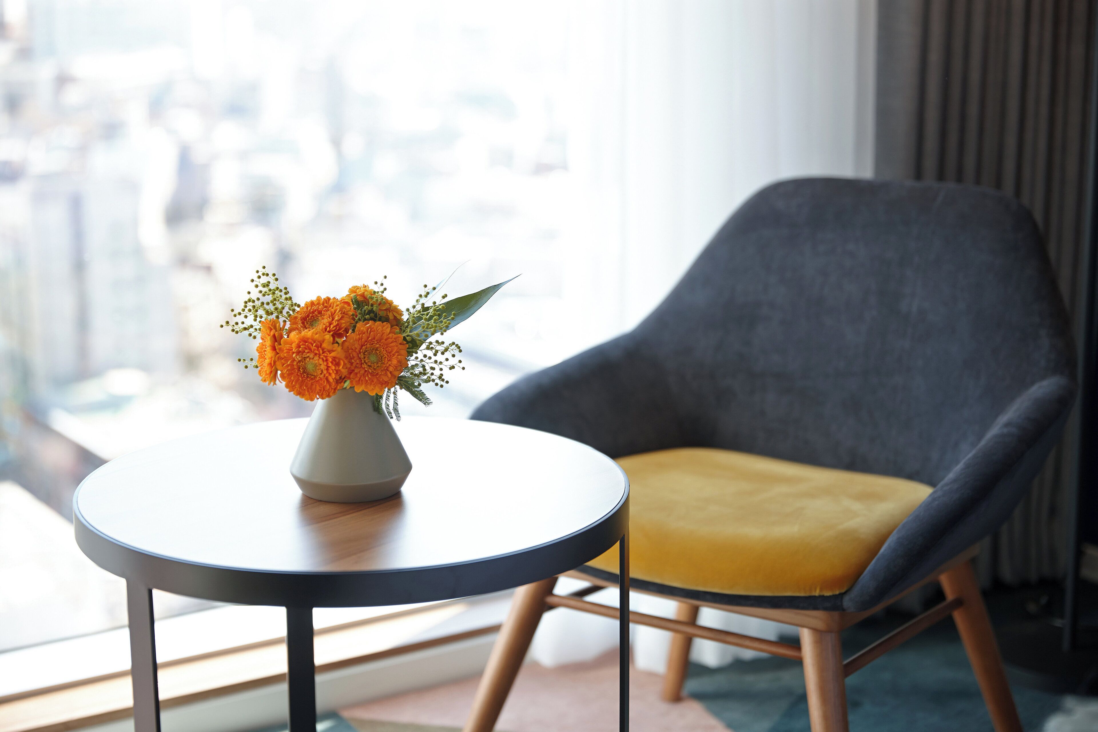 A modern room with a round table, a grey armchair with a yellow cushion, and a vase of orange flowers.