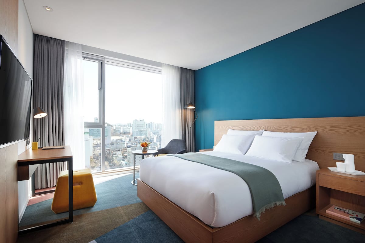 A modern hotel room with a large bed, a desk, a chair, and a view of the city.