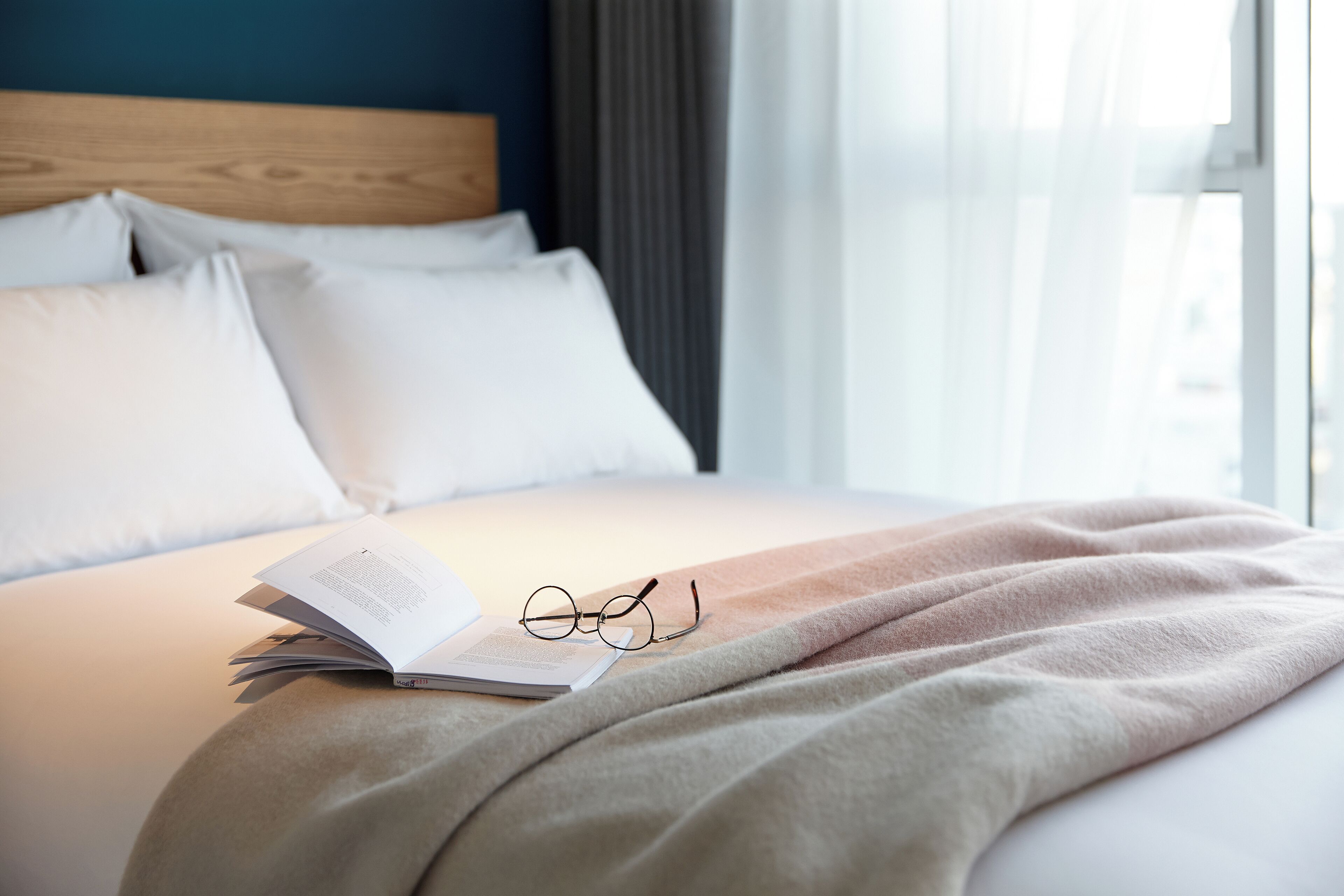 A neatly made bed with white pillows, a beige blanket, and a pair of glasses on an open book.