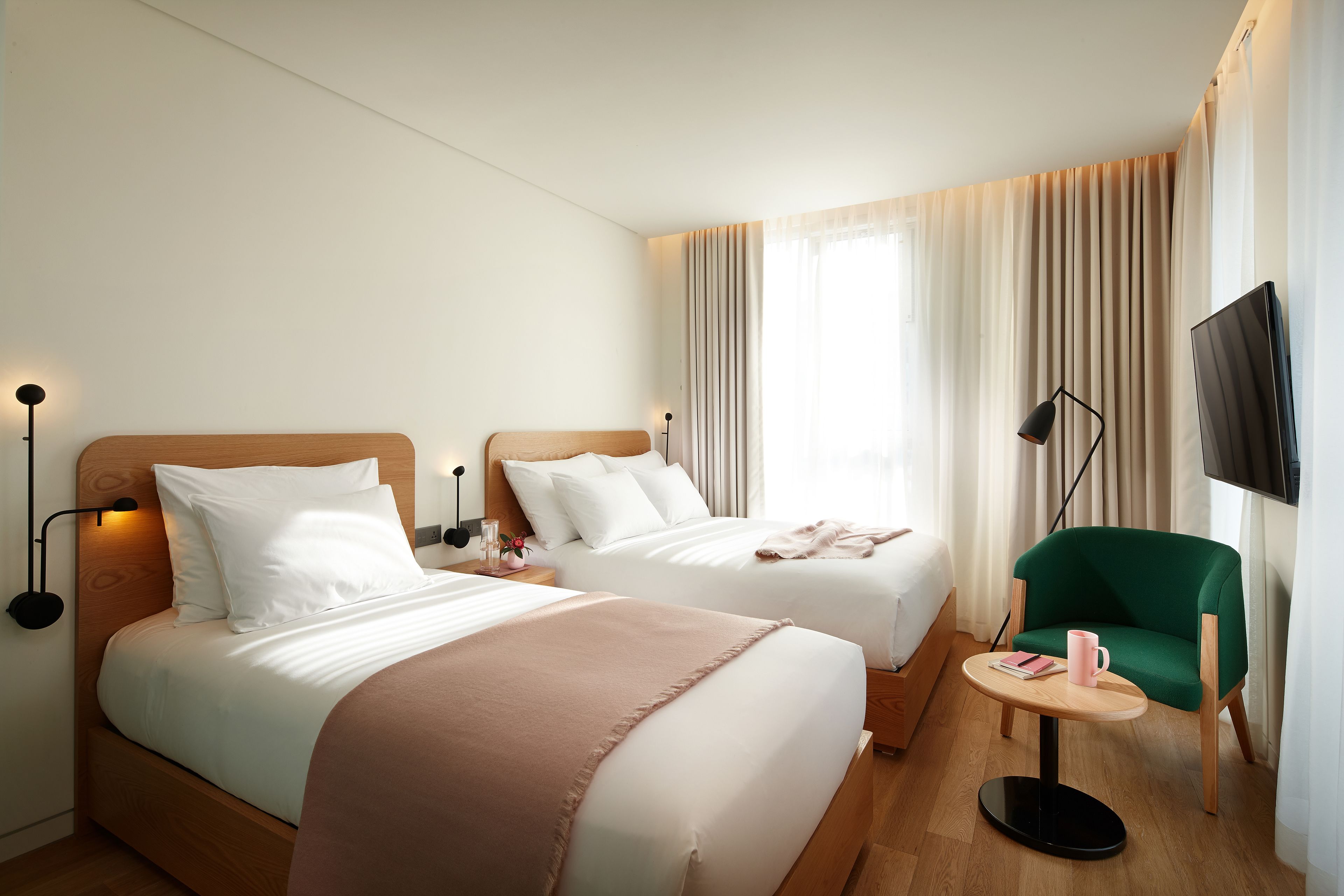 Quarto de hotel com duas camas, uma mesa redonda pequena, uma poltrona verde, televisão e uma janela com cortinas.
