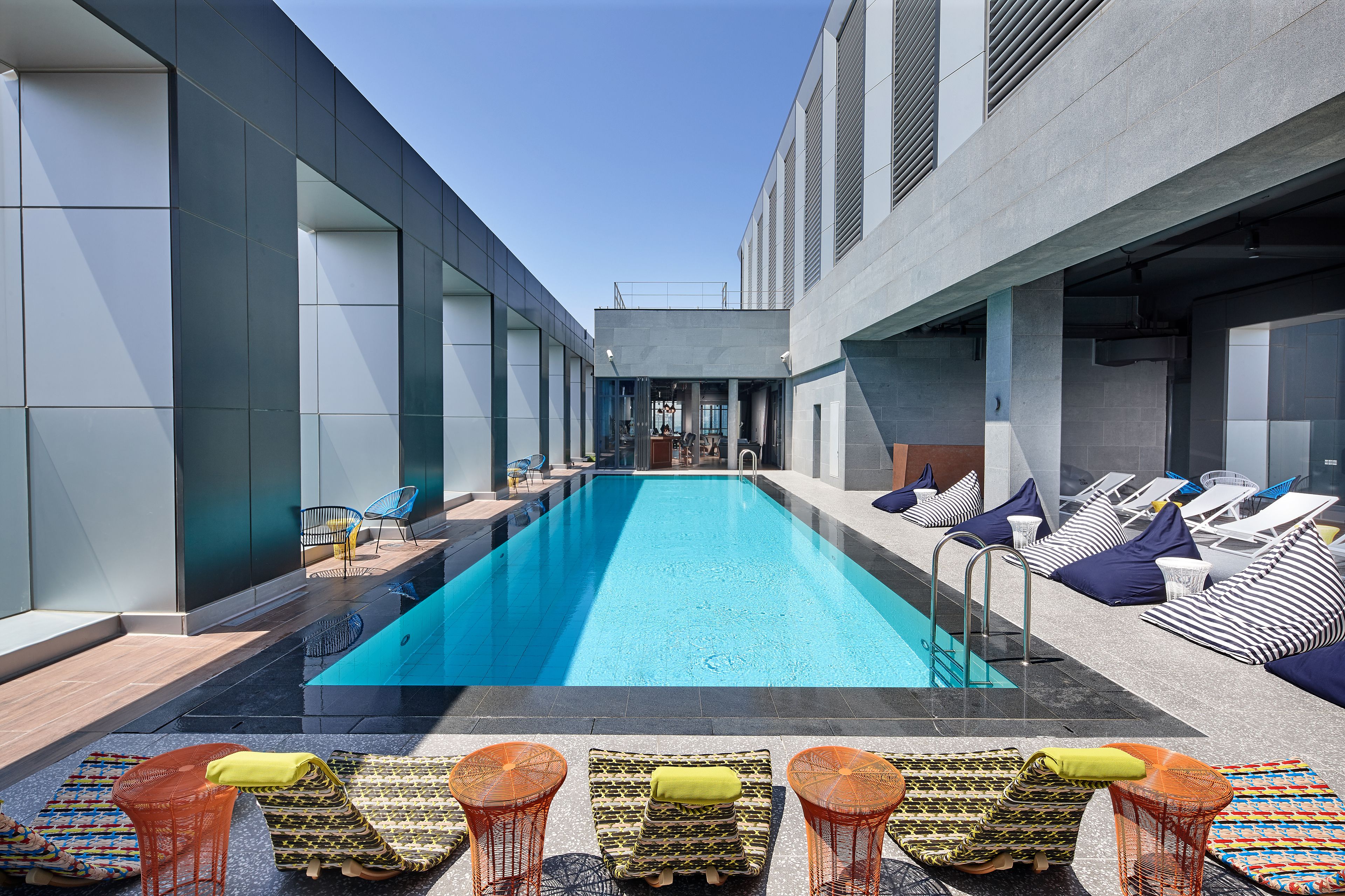 A modern pool area with lounge chairs and umbrellas, surrounded by a building with large windows and a clear blue sky.