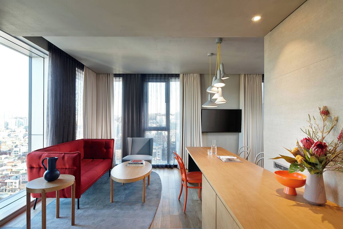 A modern hotel room with a red sofa, a wooden dining table, a small round table, and a kitchen area with a wooden countertop and orange bowl.