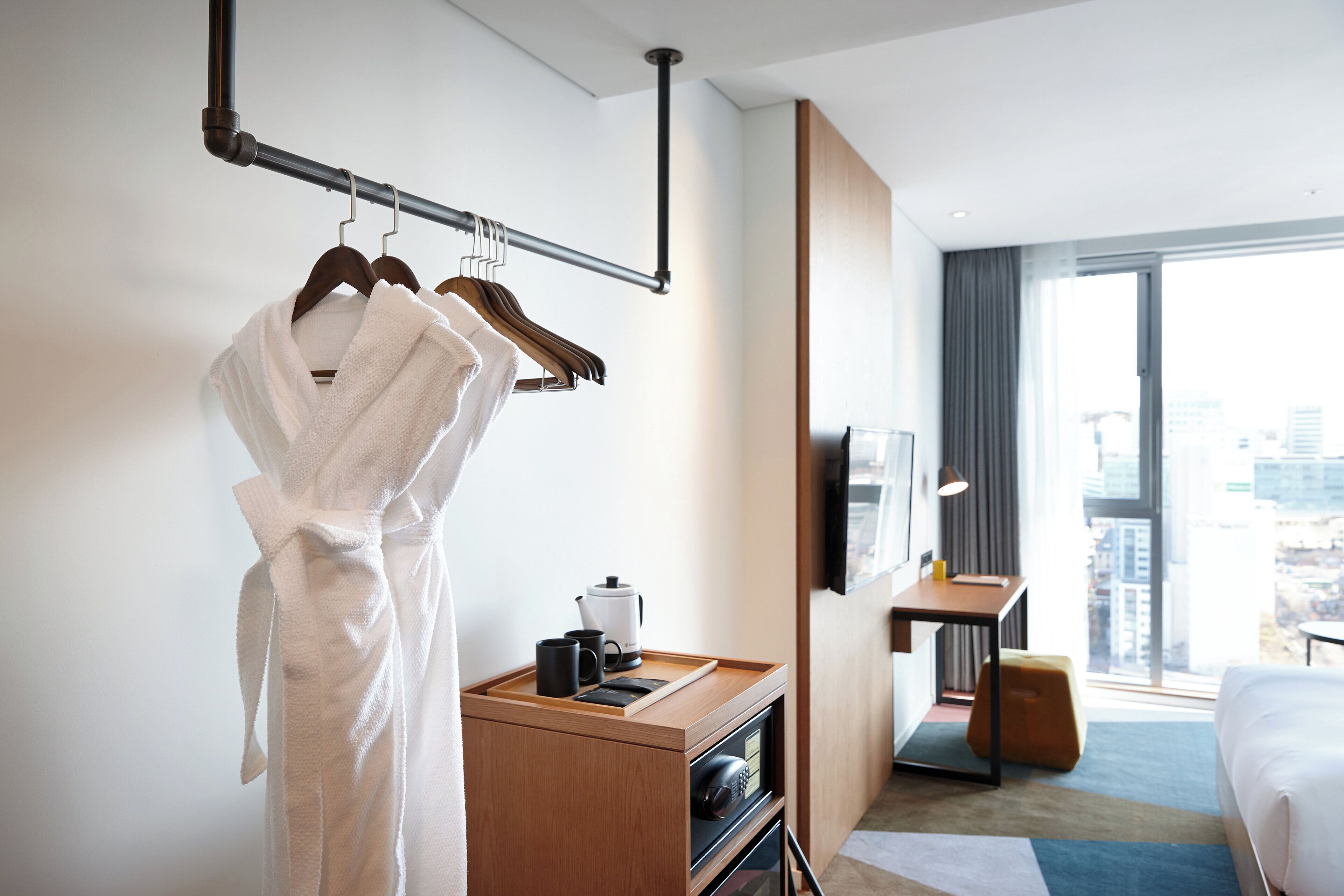 Una habitación de hotel con un escritorio de madera, una cama, una bata colgada en un perchero y vistas a la ciudad a través de la ventana.
