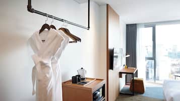 Una habitación de hotel con un escritorio de madera, una cama, una bata colgada en un perchero y vistas a la ciudad a través de la ventana.