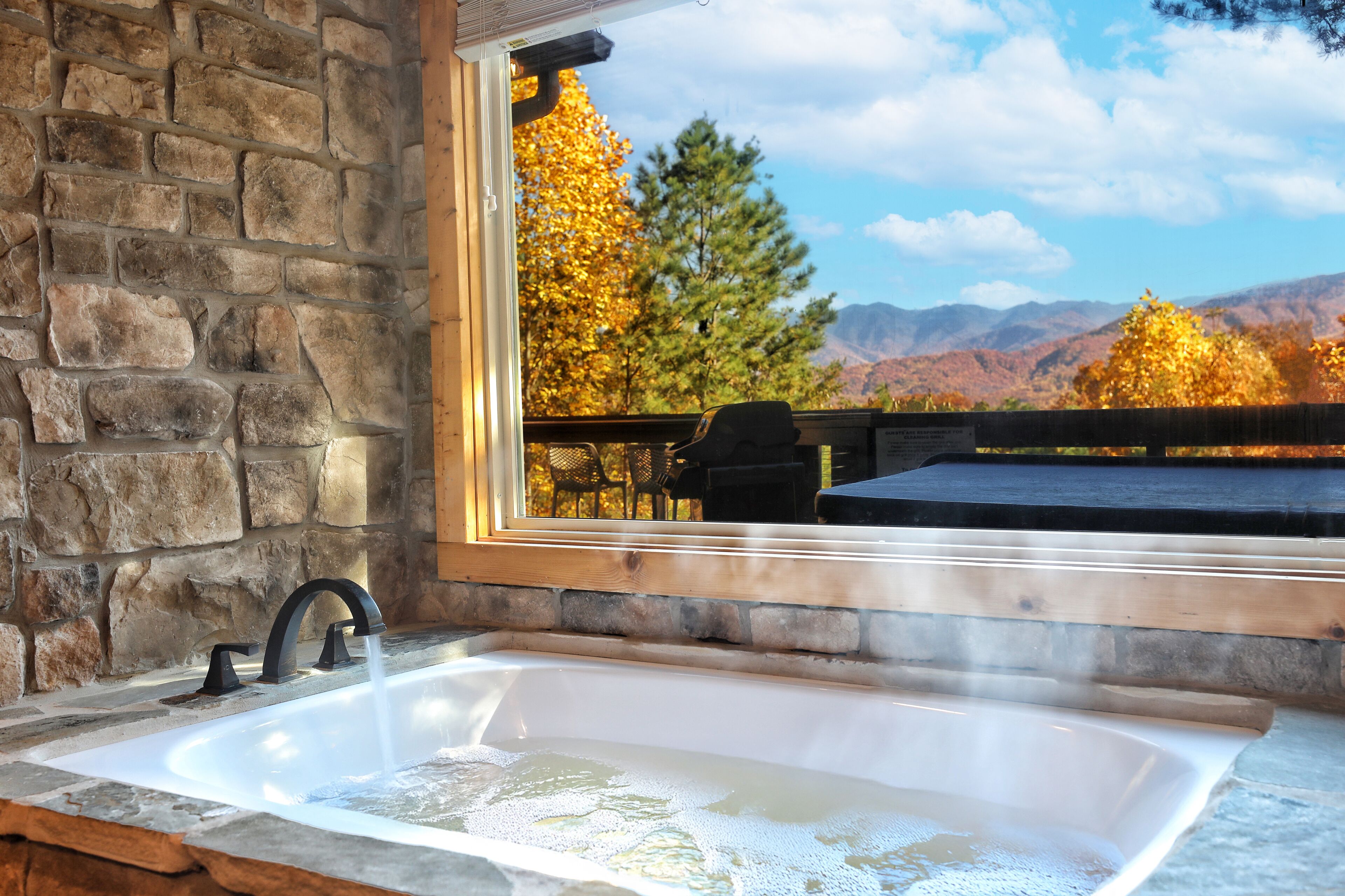 A stone bathroom with a large window, a bathtub, and a view of autumn trees and mountains.