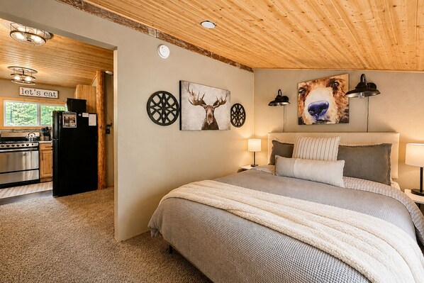 Une chambre à coucher avec un lit, deux lampes fixées au mur, un tableau représentant un chien, et une cuisine visible à travers une porte ouverte.