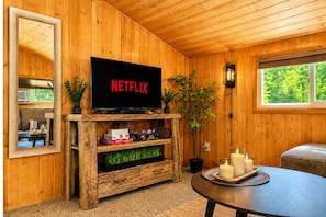 Une pièce chaleureuse avec un meuble TV en bois, un téléviseur à écran plat affichant le logo « NETFLIX », une plante en pot, des bougies sur un plateau et une fenêtre donnant sur un paysage verdoyant.