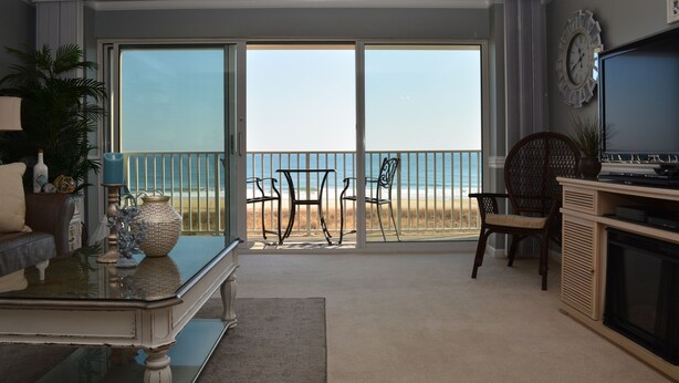 Un salon doté d’une grande porte-fenêtre coulissante donnant sur un balcon avec vue sur la plage. On y trouve un canapé, une table basse agrémentée d’objets décoratifs et un téléviseur posé sur un meuble.