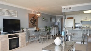 Un salon comprenant un téléviseur, une cheminée, une table à manger et des chaises. Il y a également un coin cuisine avec un réfrigérateur et un four à micro-ondes.