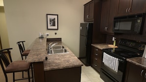 A kitchen with a granite countertop, stainless steel sink, black refrigerator, stove, and microwave. There are two bar stools and a framed picture on the wall. - Fantastic Fernie Flat !! Central Location & Amenities  (Fernie)