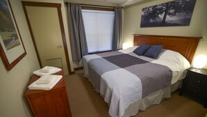 A bedroom with a large bed, a wooden nightstand, a bedside table with towels, a window with blinds, and a painting on the wall. - Fantastic Fernie Flat !! Central Location & Amenities  (Fernie)
