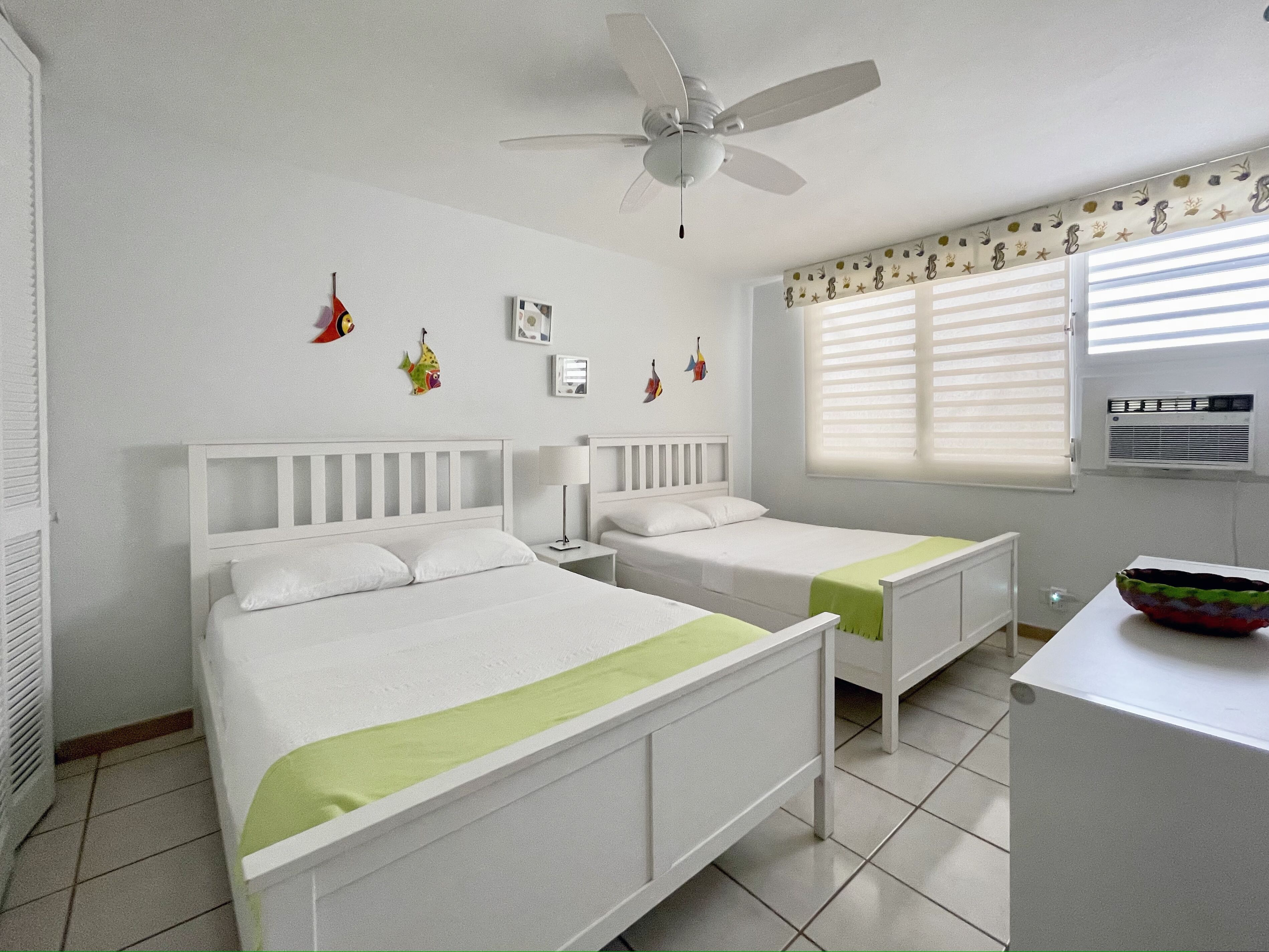 Un dormitorio con dos camas, ventilador de techo, ventana con persianas y radiador.