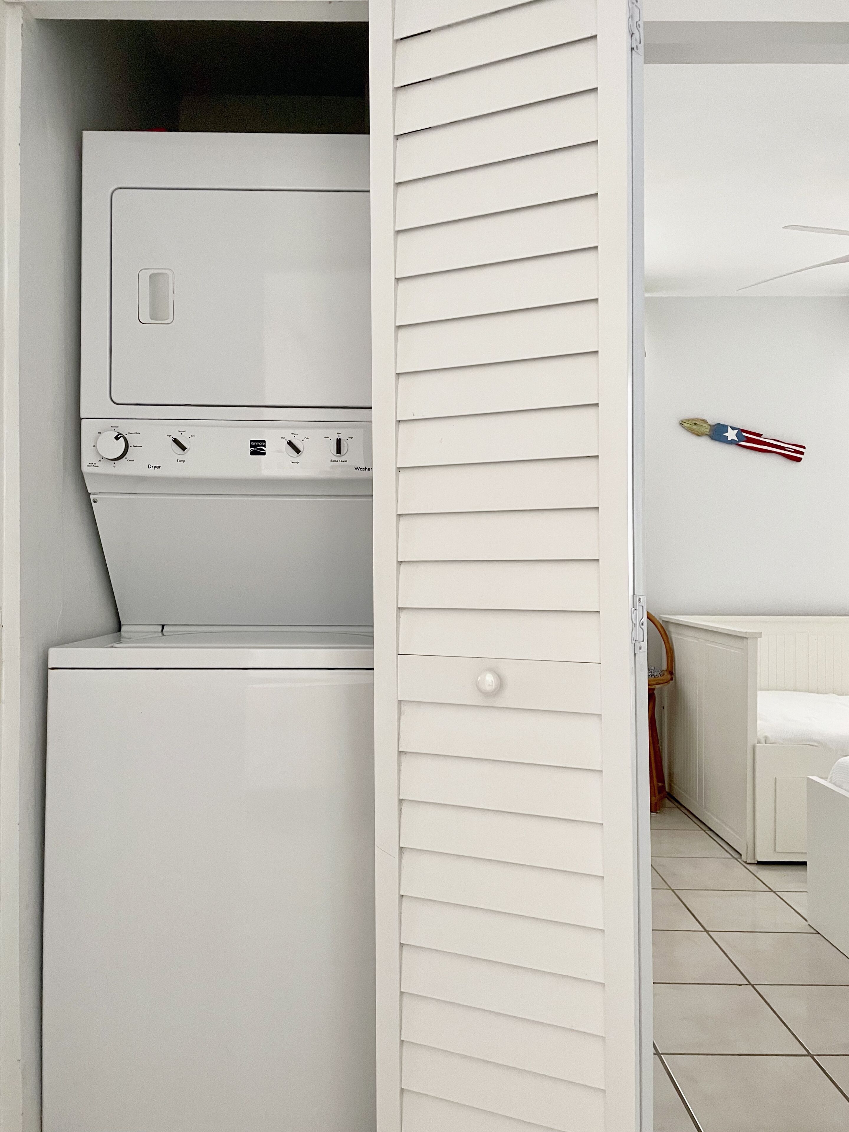 Una sala de lavandería con lavadora y secadora de carga frontal, una puerta blanca con listones horizontales y una pequeña decoración con la bandera estadounidense en la pared.