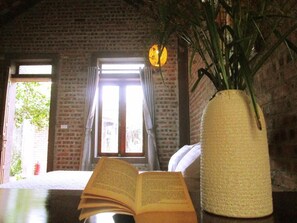 Een kamer met een bakstenen muur, een raam met gordijnen, een tafel met een open boek en een vaas met een plant.