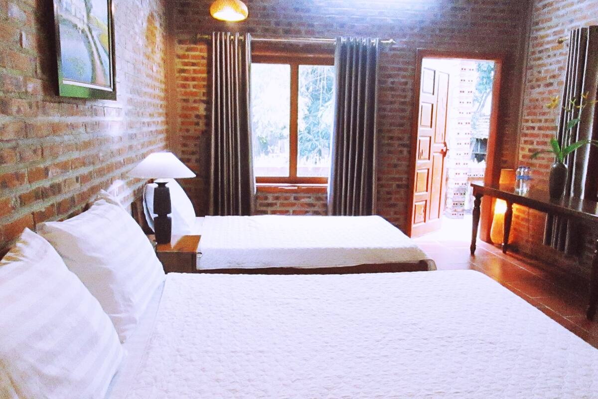 A hotel room with a large bed, a bedside table with a lamp, a window with curtains, and a brick wall.