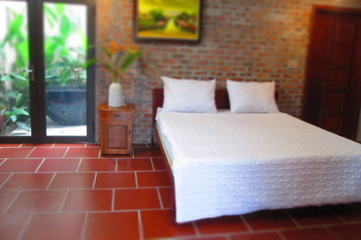 A bedroom with a brick wall, a bed with white bedding, a nightstand with a vase, and a window with greenery outside.