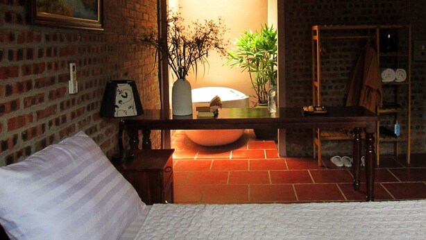 Een kamer met een bakstenen muur, een bed, een houten bankje, een spiegel, een wastafel en een plant.
