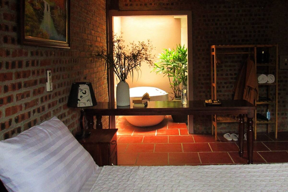 A room with a brick wall, a bed, a wooden bench, a mirror, a sink, and a plant.