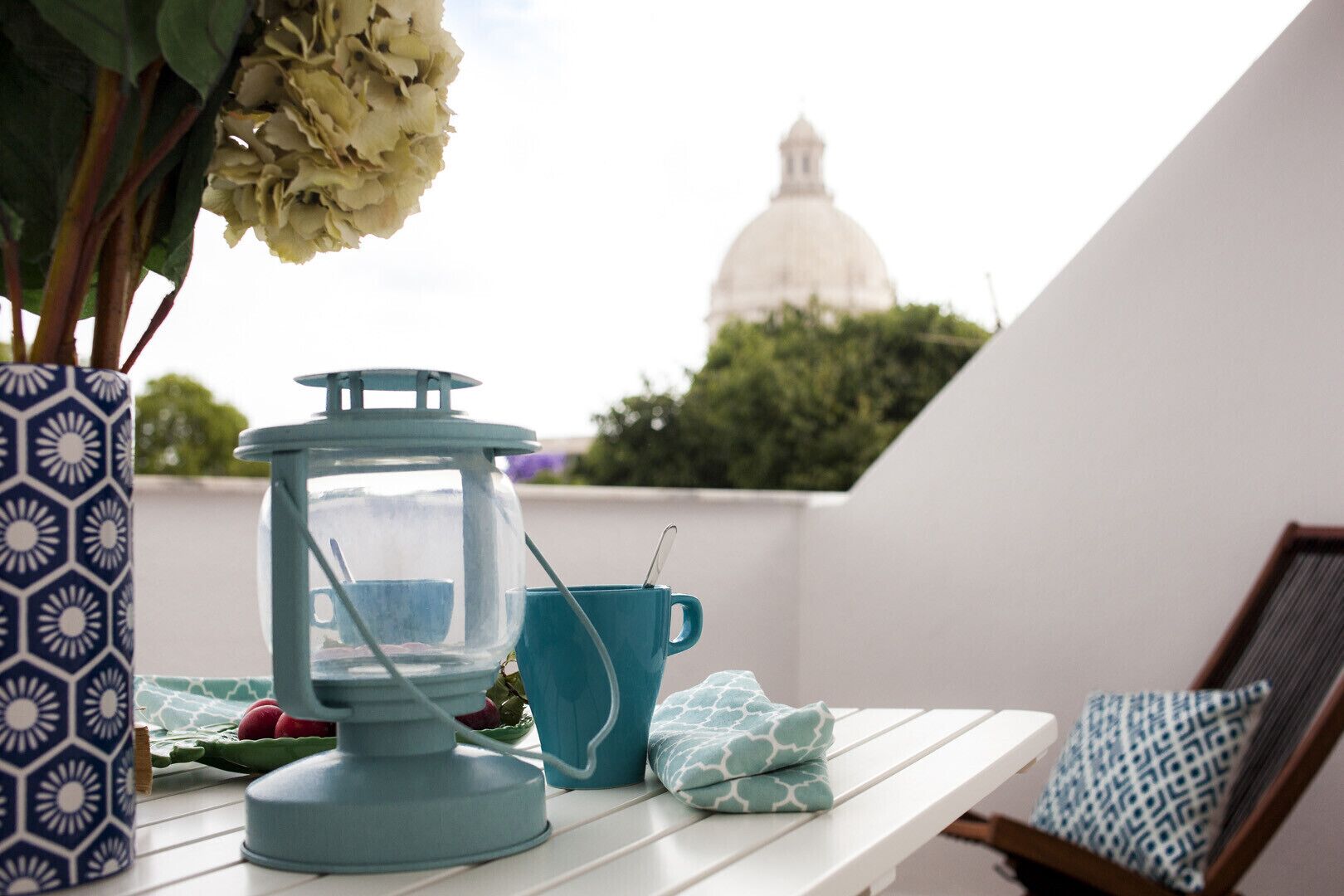 Uma mesa com um candeeiro, um vaso estampado e um almofada numa varanda com vista para um edifício em forma de cúpula.